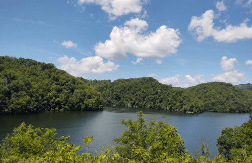 A serene lake surrounded by lush green hills under a bright blue sky with fluffy white clouds.