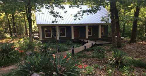 A cozy cabin with a metal roof surrounded by trees and greenery, featuring a wooden walkway and colorful plants.