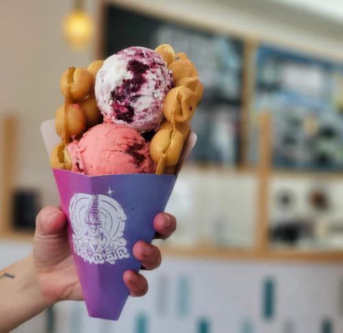 A hand holding a colorful ice cream cone with three scoops and waffle balls, set against a blurred café background.