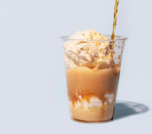 A clear cup filled with ice cream and whipped cream, with a brown liquid being poured on top.