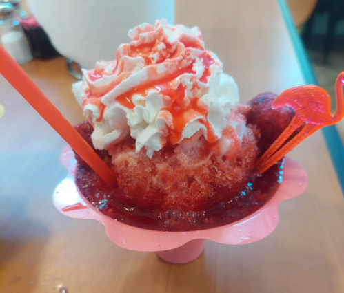 A colorful snow cone topped with whipped cream, syrup, and decorative flamingo straws in a pink cup.