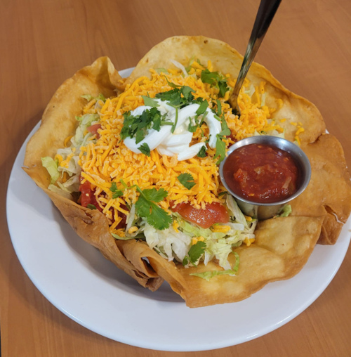 A crispy tortilla bowl filled with lettuce, tomatoes, cheese, cilantro, topped with sour cream and served with salsa.