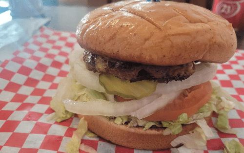 A juicy hamburger with lettuce, tomato, pickles, and onions on a toasted bun, served on a red and white checkered paper.