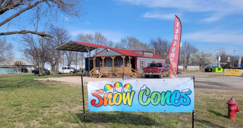 A colorful "Snow Cones" banner in front of a small building with a porch, truck, and trees in a grassy area.