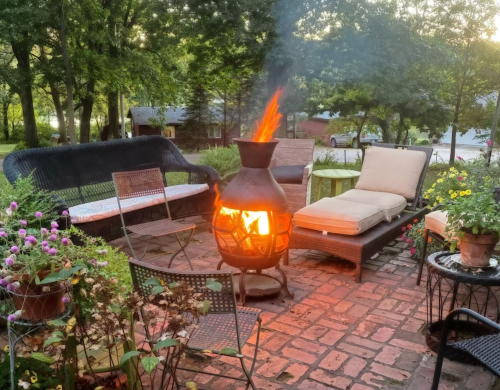 A cozy outdoor seating area with a chiminea fire, surrounded by greenery and colorful flowers.