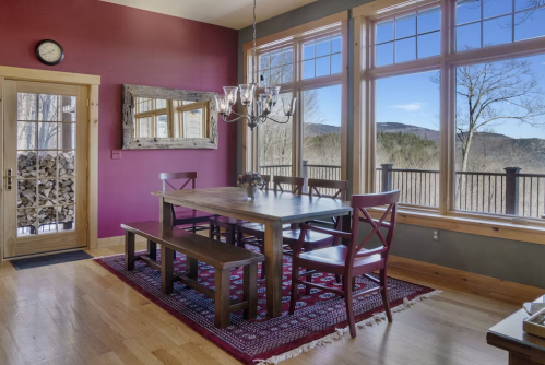 Cozy dining area with a wooden table, red chairs, large windows, and mountain views. Warm, inviting atmosphere.