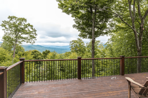 A wooden deck with black railings overlooks a lush green landscape and distant mountains under a cloudy sky.