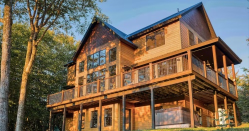 A large wooden house with multiple windows and a deck, surrounded by trees and bathed in warm sunlight.