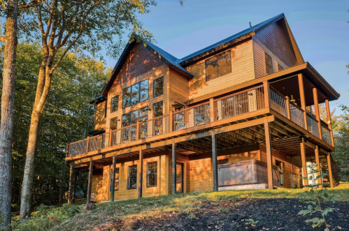 A large wooden house with multiple windows and a spacious deck, surrounded by trees and bathed in warm sunlight.