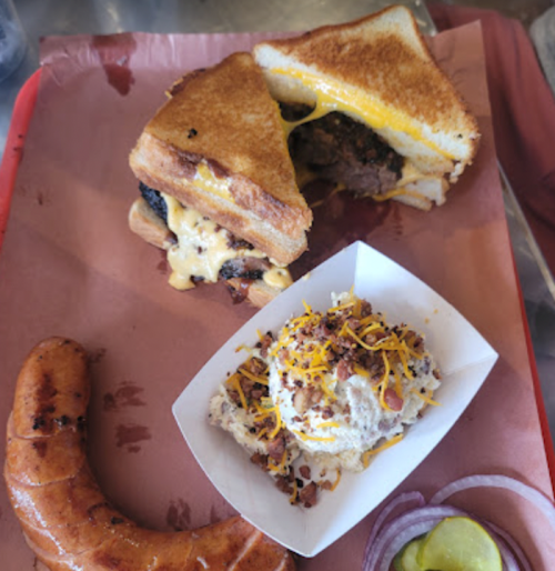 A grilled cheese sandwich with meat, a side of creamy potato salad topped with bacon and cheese, and a sausage link.