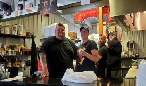A group of four people smiling behind a food counter, with various kitchen items and a "Pick Up Here" sign visible.