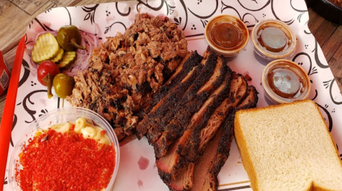 A tray with sliced smoked brisket, pulled pork, macaroni salad, pickles, and bread, accompanied by barbecue sauce.