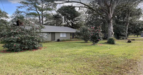A single-story house surrounded by green grass and trees, with flowering bushes in the front yard.
