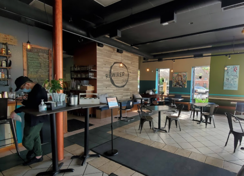 A cozy café interior with tables, a counter, and a barista preparing drinks. Brightly lit with colorful decor.
