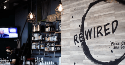 Interior of a modern pizza cafe named "Rewired," featuring a wooden wall sign and a bar area in the background.