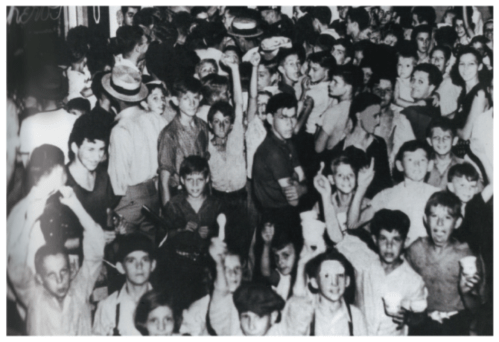 A crowded black-and-white scene of a large group of people, mostly children and young adults, in a lively setting.