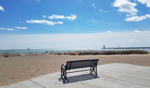A black bench on a sandy beach overlooking a calm lake under a blue sky with scattered clouds.