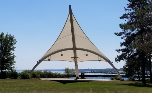 A large, white sail-like structure stands in a park by the water, surrounded by trees and greenery.