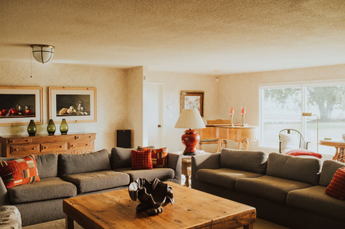 Cozy living room with gray sofas, wooden coffee table, and large windows, featuring warm decor and natural light.