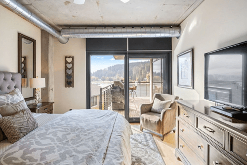 Cozy bedroom with a large window, modern decor, and a view of nature outside. Features a bed, armchair, and TV.