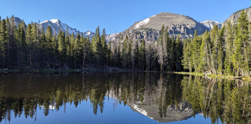 A serene mountain lake reflecting surrounding trees and peaks under a clear blue sky.