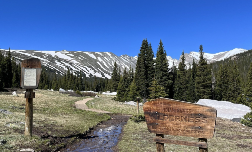 Trailhead sign for Indian Peaks Wilderness, surrounded by snow-capped mountains and pine trees under a clear blue sky.