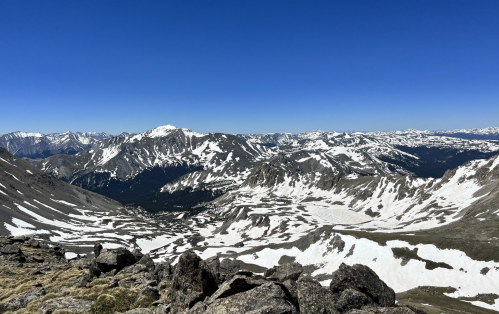 A panoramic view of snow-capped mountains under a clear blue sky, with rocky terrain in the foreground.