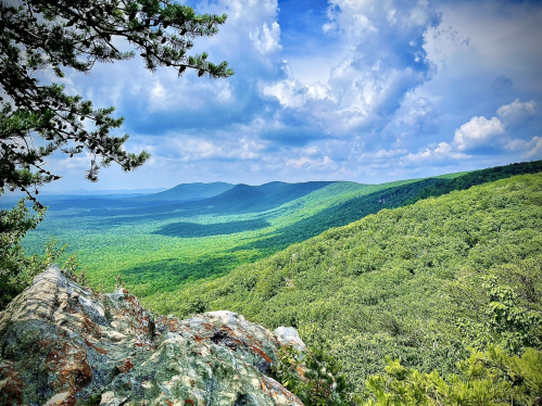 A panoramic view of lush green mountains under a partly cloudy sky, seen from a rocky overlook.