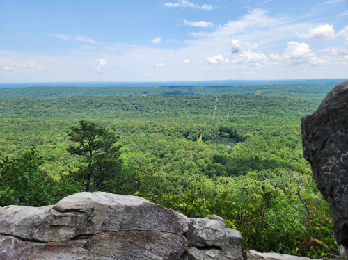 A panoramic view of lush green forests and a distant lake, framed by rocky outcrops under a blue sky with clouds.