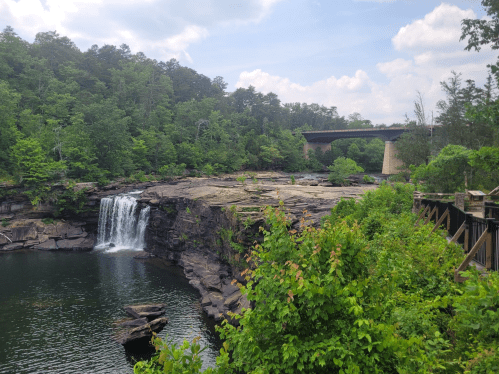 A serene waterfall cascades into a calm pool, surrounded by lush greenery and a bridge in the background.