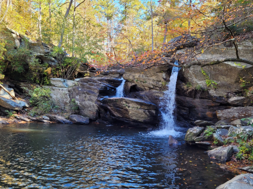 A serene waterfall cascades over rocks into a calm pool, surrounded by autumn foliage and trees.