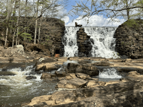 A waterfall cascades over a stone dam, surrounded by trees and rocky terrain under a partly cloudy sky.