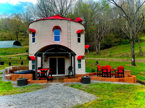 A unique round house with a red roof, surrounded by greenery and outdoor seating on a gravel path.