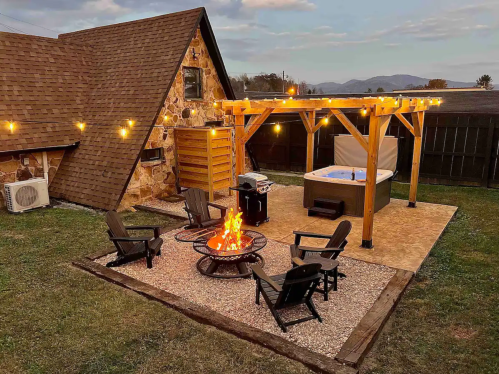 Cozy outdoor space with a fire pit, seating, hot tub, and string lights near a rustic stone house.