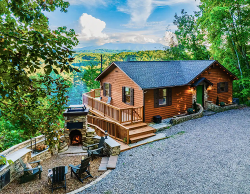A cozy wooden cabin surrounded by trees, featuring a deck, outdoor seating, and a scenic view of the mountains.