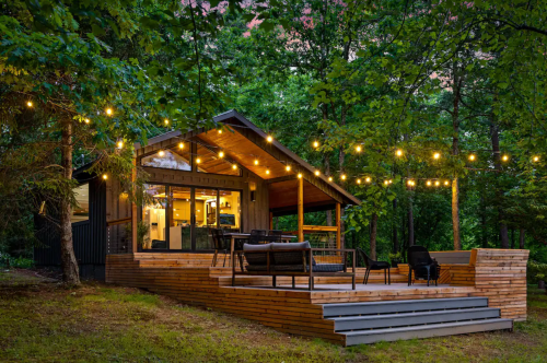 A cozy cabin in the woods, featuring string lights, a wooden deck, and surrounded by lush greenery.