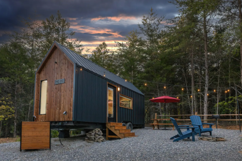 A cozy tiny house on wheels surrounded by trees, with a deck, chairs, and string lights under a dramatic sunset sky.