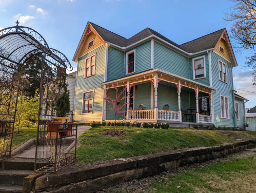 A charming blue Victorian house with a porch, surrounded by greenery and a decorative archway in the foreground.