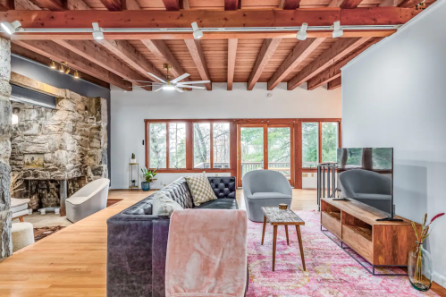 Cozy living room with wooden beams, stone fireplace, modern furniture, and large windows overlooking greenery.