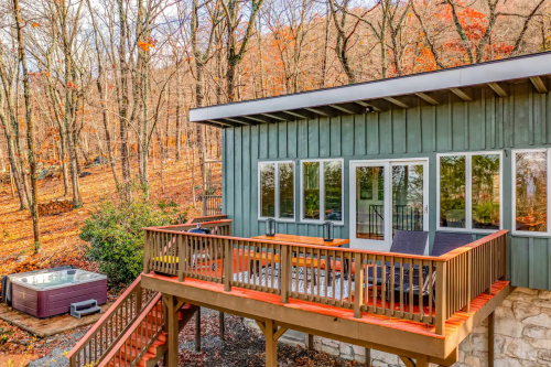 A modern cabin with a deck and hot tub, surrounded by autumn trees and a scenic hillside.