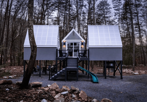 A modern cabin with a unique design, featuring large windows, a slide, and surrounded by trees in a wooded area.