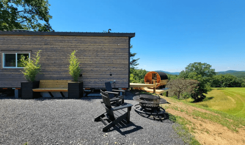 A cozy tiny home with outdoor seating and a fire pit, set against a scenic mountain backdrop on a sunny day.