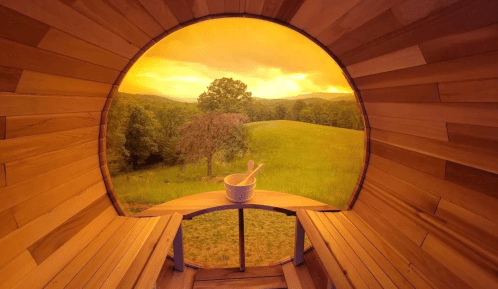 A circular wooden room with benches overlooks a scenic landscape at sunset, featuring rolling hills and a tree.