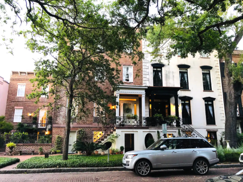 A historic building with a mix of brick and white facade, surrounded by trees, with a parked silver SUV in front.