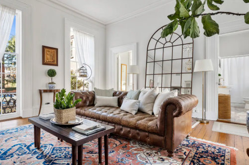 Cozy living room with a leather sofa, decorative pillows, a coffee table, and large windows letting in natural light.