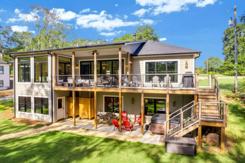A modern two-story house with a spacious deck, outdoor seating, and a hot tub, surrounded by greenery and blue skies.