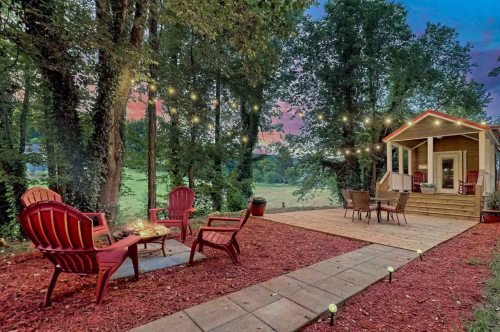 Cozy outdoor patio with red chairs, a fire pit, and string lights, surrounded by trees and a scenic view at sunset.