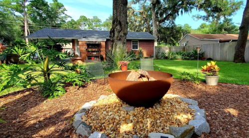 A backyard scene featuring a fire pit surrounded by stones, plants, and a cozy home in the background.