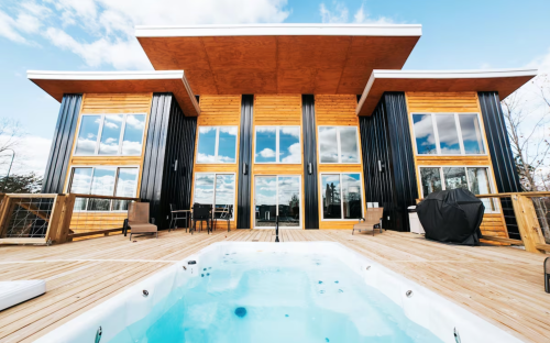 Modern wooden house with large windows, a deck, and a hot tub in the foreground under a blue sky with clouds.
