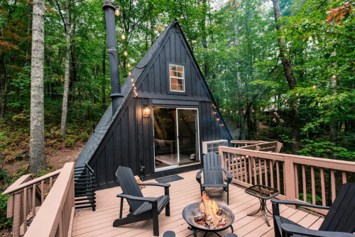 A cozy A-frame cabin surrounded by trees, featuring a deck with chairs and a fire pit.